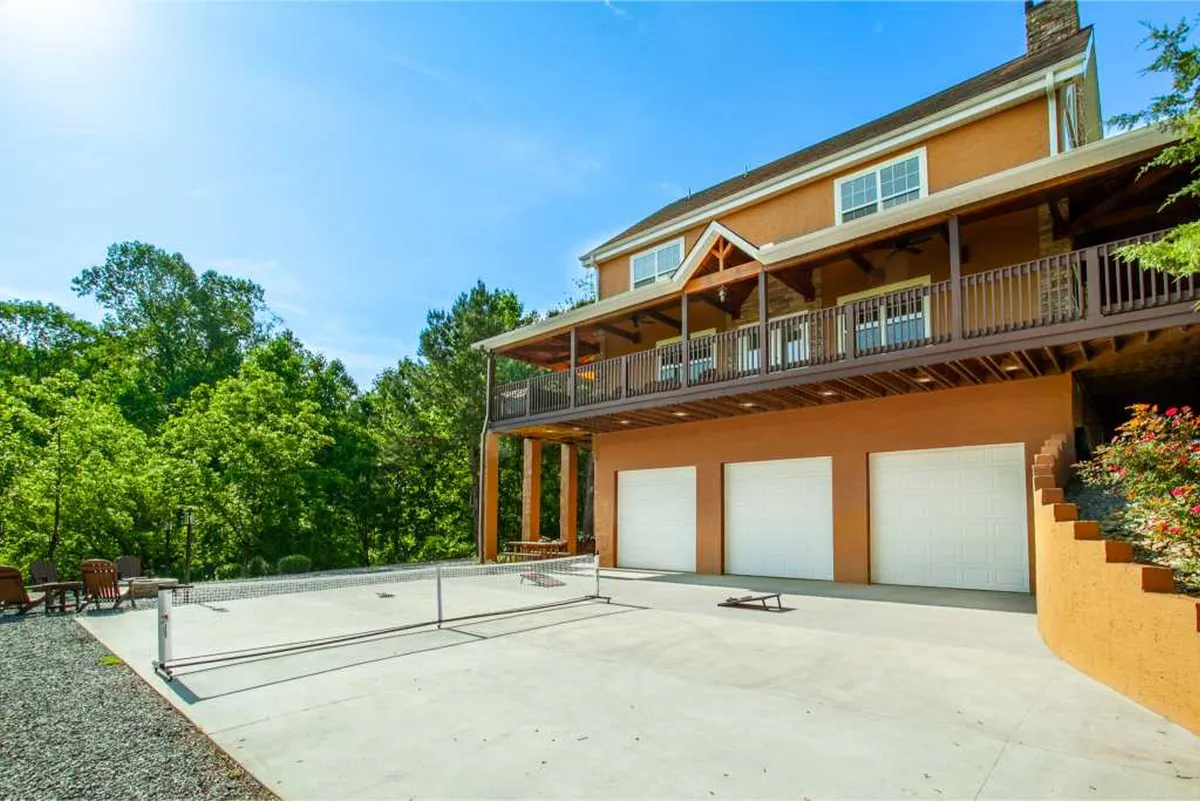A spacious exterior view of the large house featuring a balcony, garage doors, outdoor seating area, and surrounded by lush greenery. The patio area suggests space for gatherings, relaxation, and sports activities, all under sunny skies.