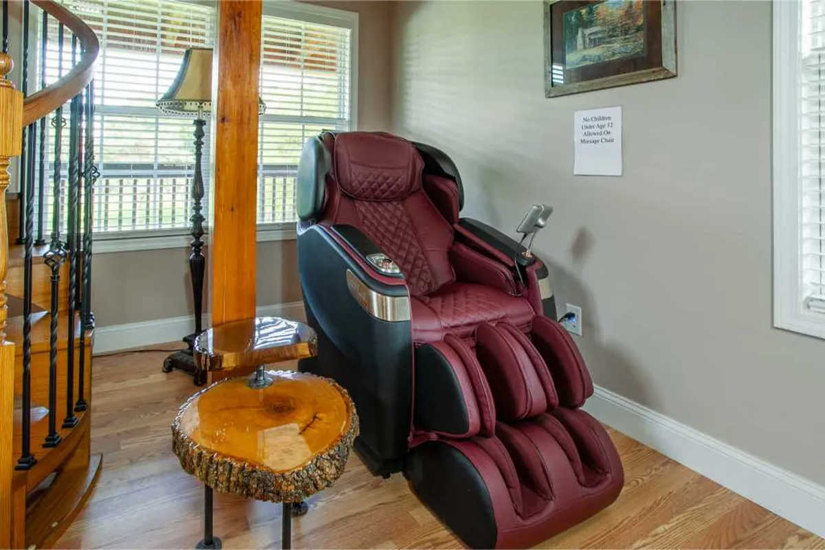 An inviting interior space with a modern massage chair set beside a wooden side table and a spiral staircase. A window lets in natural light, and a cozy lamp adds a relaxing ambiance.