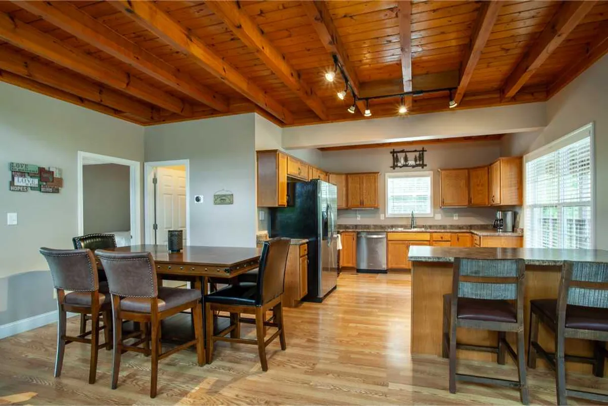 A cozy interior with a central dining table, a kitchen island, exposed wooden beams, and modern appliances. Windows in the background hint at a connection to nature and fill the room with soft, natural light.