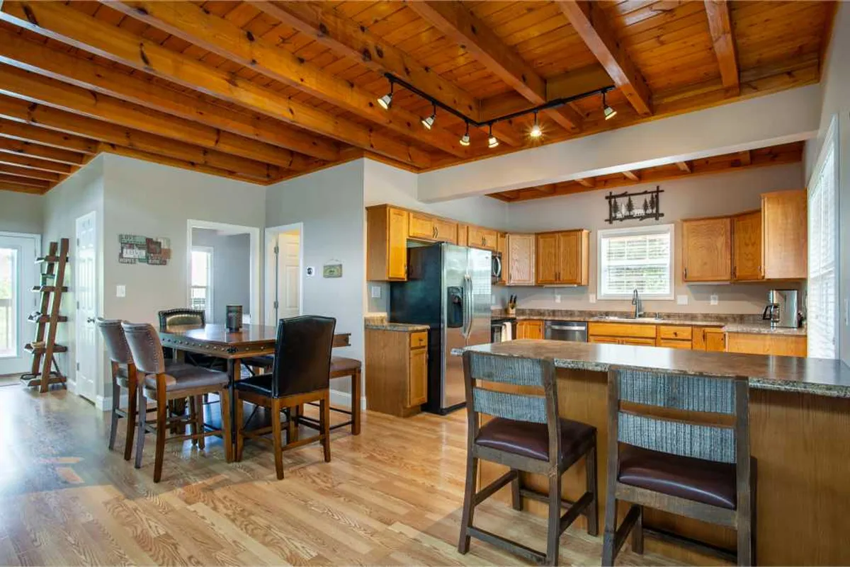 A warm, welcoming kitchen and dining space with wooden beams overhead, a prominent kitchen island, a large dining table, and sleek stainless steel appliances. The layout encourages easy movement and connection between cooking and dining activities.