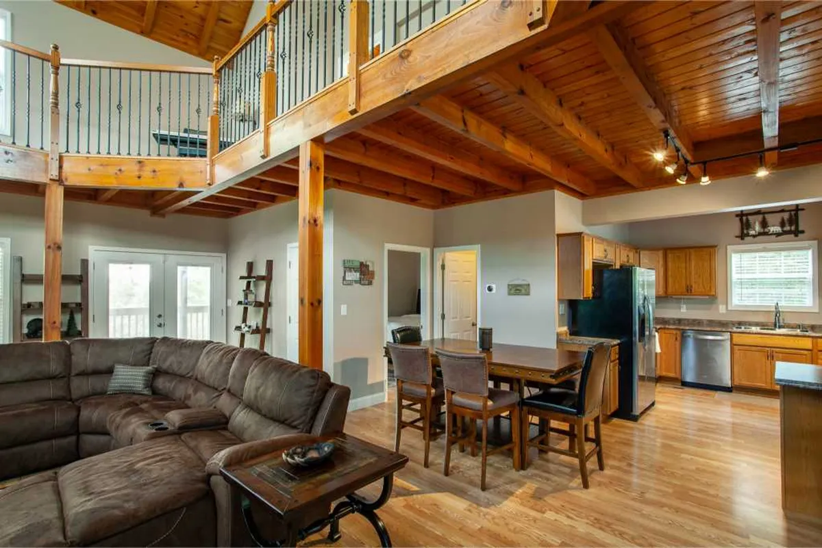 A spacious open-concept living area featuring a large sectional sofa, a generous dining table set for a group, and a modern, fully equipped kitchen in the background. The space is bathed in natural light from expansive windows, creating an inviting and communal atmosphere ideal for family meals and gatherings.