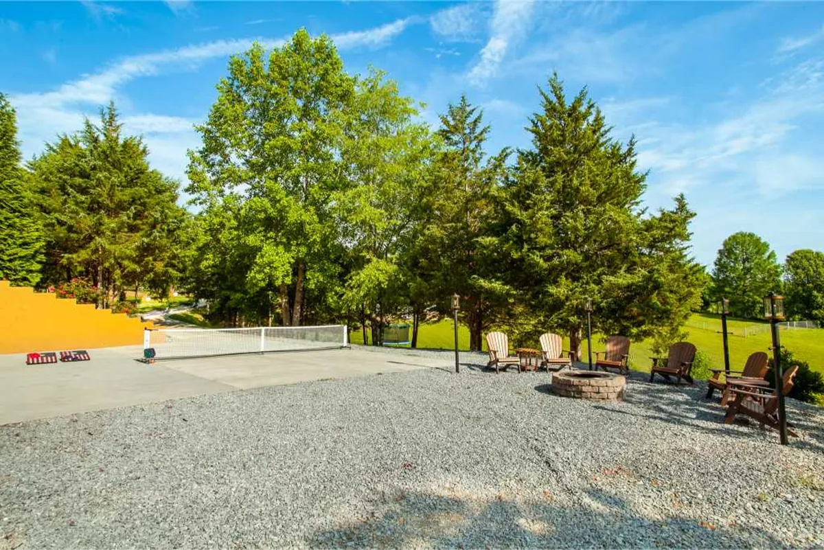 The image shows a gravel patio with a cozy fire pit encircled by Adirondack chairs, all framed by lush green trees and a striking orange wall. The inviting space is perfect for evening gatherings, stargazing, or intimate conversations by the fire.
