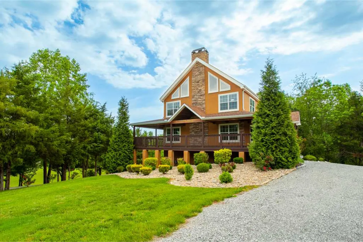 The hero image showcases a charming single-family house with a welcoming wraparound porch, beautifully landscaped garden, and spacious gravel driveway. The scene captures the inviting warmth of the home at sunset, with soft golden light illuminating the porch—perfect for relaxing mornings with coffee or winding down after a day of adventure. The expansive yard hints at plenty of space for family fun and outdoor activities.